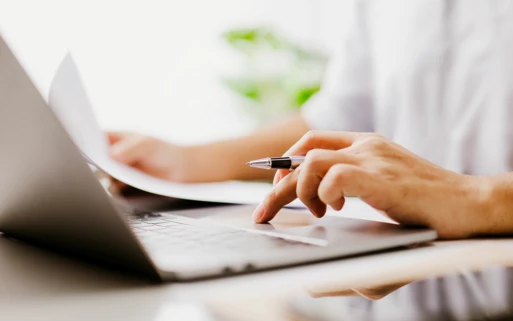 Nahaufnahme einer Person, die Dokumente überprüft und auf einem Laptop arbeitet